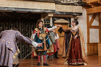 The Duke in the center of the stage with long curly brown wig, a colorful mesh of robes, a staff in his right hand and his left hand holding high the and of Silvia in a burgundy and gold Jacobean dress and gold cape as, in the front left corner, Valentine bows, his light purple coat's back to us.
