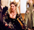 Livia in black dress with floral sleeves and skirt and gold lace high collar screams as she kneels on the floor, other characters standing in the background