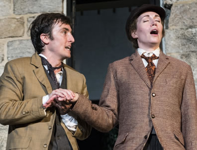 Orlando in brown jacket and olive vest holds Gannymede's right hand in his two hands; she is wearing a brown checked jacket with orange-black-patterned necktie and black bowler hat, her eyes closed as she speaks, and he looks like he's trying to appease her. Stone walls in background.