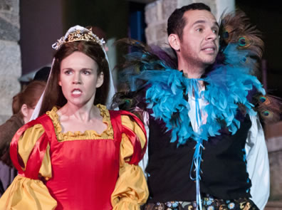 Kate in red Elizabethan dress with gold sleeves and trim, a crown and white head dress; Petrucio in black vest, white shirt, multi-colored boxer shorts, and a blue boa collar with peacock feathers