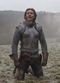 Henry in armor on his knees on the top of a trampled meadow hill with his sword stuck in the ground nearby, and the soldier's gage in his belt