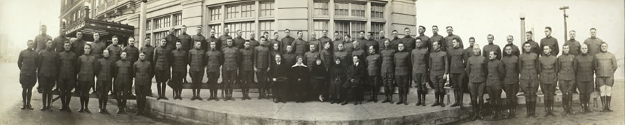 World War I U.S. Army soldiers in three rows with five people in formal suits and dresses sitting in a line in the middle, with a hotel facade in the background.