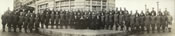 World War I U.S. Army soldiers in three rows with five people in formal suits and dresses sitting in a line in the middle, with a hotel facade in the background.