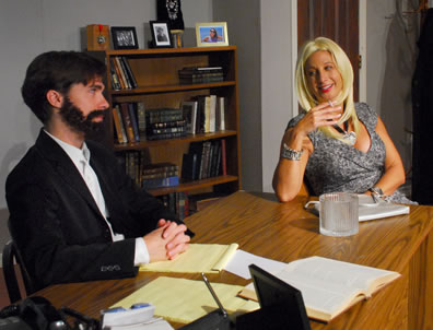 Rabbi Yoshi in black coat and beard, hands clasped on a yellow legal pad on his cluttered desk looks sideways at Ruth, smoking a cigarette, wearing a diamond bracelet and pendant and a silver pattern dress with plunging neckline; bookcase with texts, photos, and awards in background against wall and next to a door.