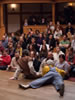 One actor sits on the edge of the stage, another lies next to him as a full audience watches in the small playhouse