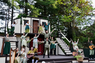 On a two-tiered white and timber stage, with stairs up each flank, the company finishes a song with arms outstretched. Trees crowd the back of the stage.