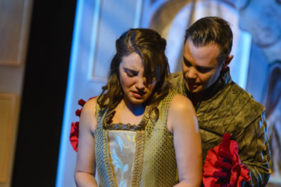 Imogen in brocaded blouse with bare arms cringes as Iachimo in green Renaissance jacket, red flowery cuffs and red gloves comes up behind her to put his hands on her shoulders.