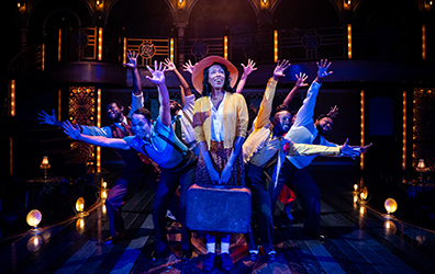 Vy, wearing a jellow jacket, white blouse, orange patterend dress, white socks, plain shoes, and a wide brim hat stands with both hands holding a battered suticase in front of her as the ensemble behind her leans out with outstreched hands creating a starburst effect. Photo by Daniel Rader