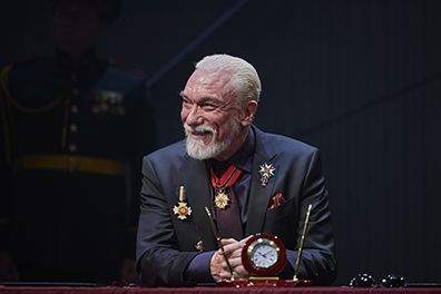 STC-Lear_Page-50037 Patrick Page as King Lear, wearing a dark blue suit with purple tie, his jacket adorend with medals and a med on a red and blue ribbon around his neck, sits at a desk, arms folded and sporting a gentle smile. In front of him on the desk is a clock-centered pen stand. Photo by DJ Corey Photography.