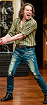 Aiden O'Reilly as Petruchio laughing in The Taming of Shrew in a vertically green striped work shirt and faded blue jeans 
