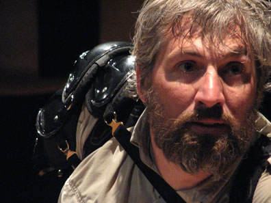 Face close to the camera on the right of the frame, brown hair and full beard, khaki jacket, and what looks like layers of a shoulder or knee pad strapped to his right shoulder